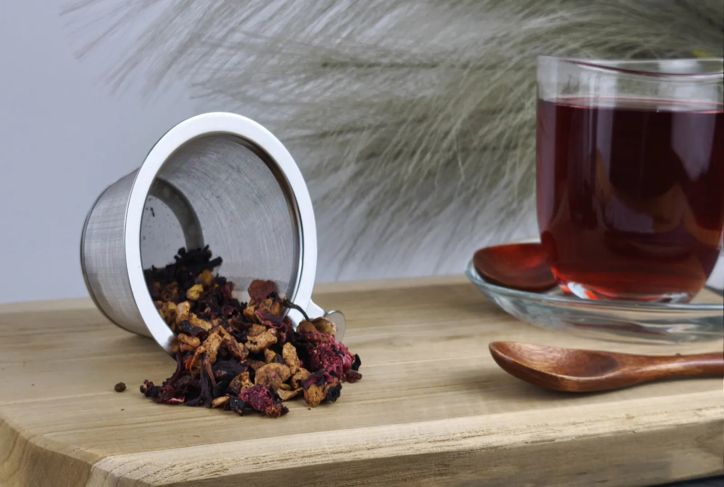 „Loser Früchtetee fällt aus einem Edelstahl-Teesieb auf ein Holzbrett, daneben steht eine Glas-Tasse mit aufgegossenem, dunkelrotem Tee und ein kleiner Holzlöffel.