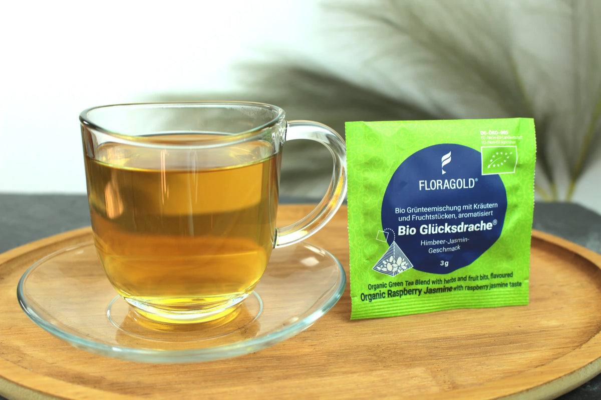 Produktfoto einer Tasse Tee und der Verpackung von Bio Glücksdrache, Teebeutel für sanften Genuss, Bio Grünteemischung mit Kräutern und Fruchtstücken, Himbeer-Jasmin-Geschmack.