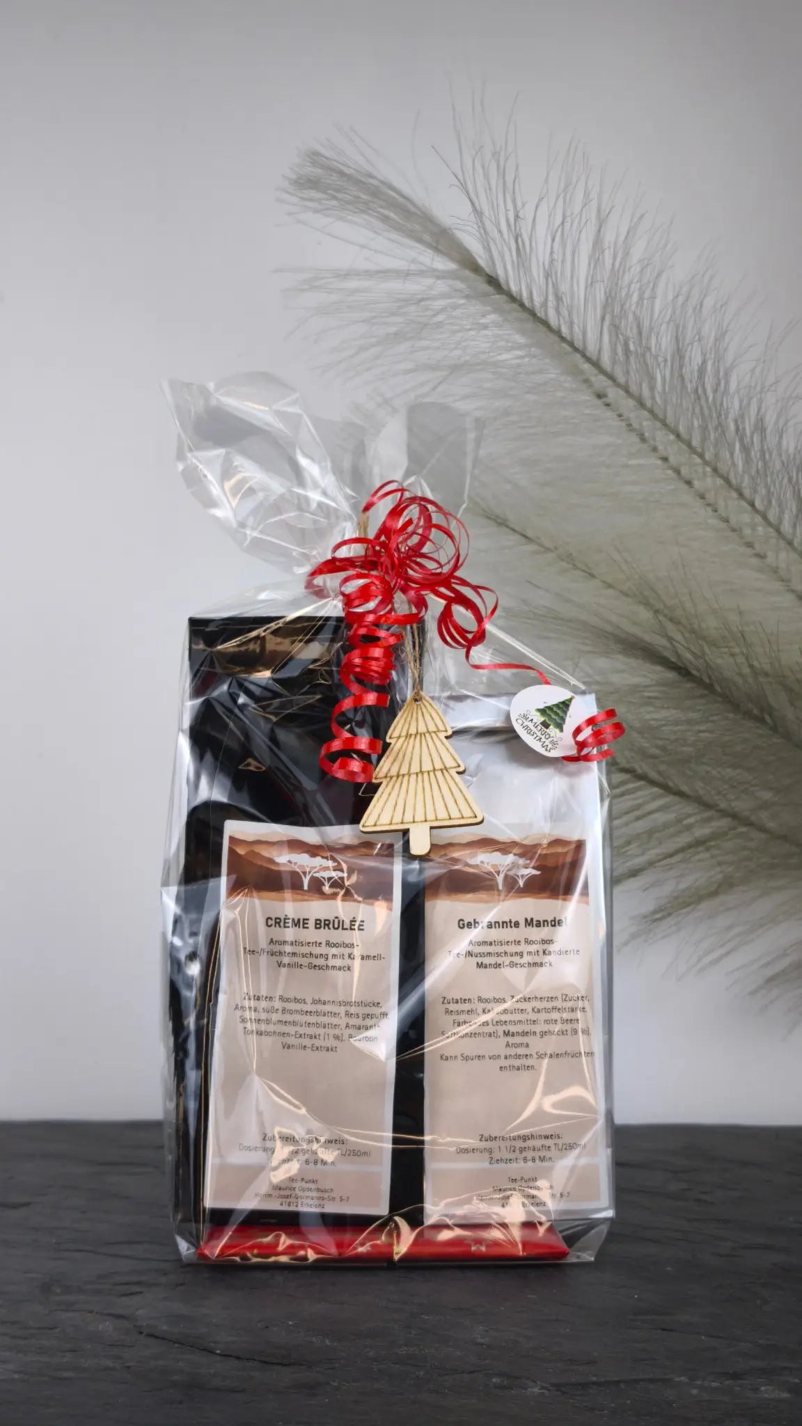 Geschenkset mit Crème Brûlée und Gebrannte Mandel Tee, festlich verpackt in Geschenkfolie mit Weihnachtsanhänger und roter Schleife.