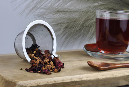„Loser Früchtetee fällt aus einem Edelstahl-Teesieb auf ein Holzbrett, daneben steht eine Glas-Tasse mit aufgegossenem, dunkelrotem Tee und ein kleiner Holzlöffel.