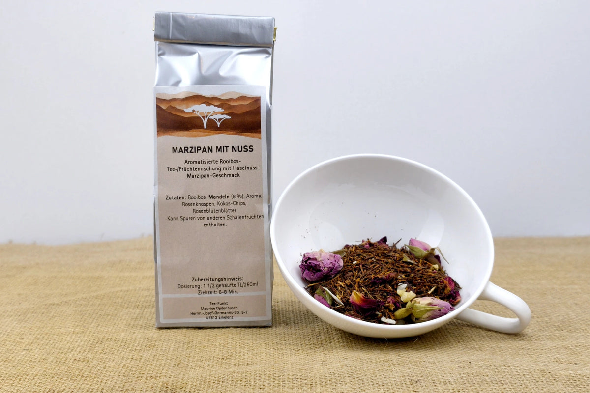 Silberne Packung Marzipan mit Nuss Rooibostee neben einer weißen Tasse mit losem Rooibostee, Rosenblüten und Nussstückchen.
