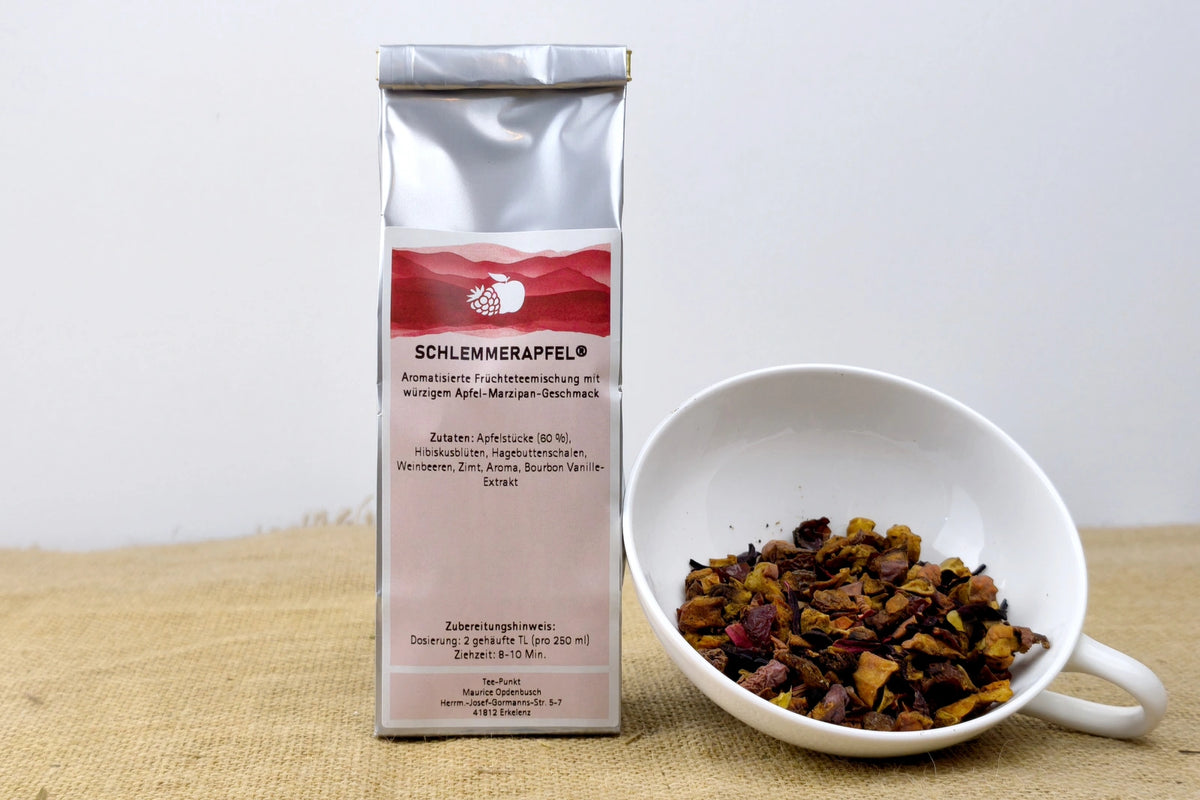 Silberne Packung Schlemmerapfel Früchtetee mit Apfel-Marzipan-Geschmack neben einer weißen Tasse mit losem Früchtetee.