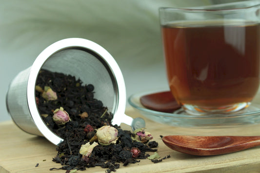 Magie des Waldes Schwarztee, loser schwarzer Tee mit Rosenknospen im Teesieb, Teetasse mit warm braunem Schwarztee-Aufguss auf Untertasse und Holzlöffel auf Holzuntergrund