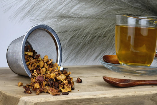 Früchtetee Passionsfrucht-Vanille, loser Tee im Edelstahl-Teesieb, goldgelber Teeaufguss in Glas, Holzlöffel auf Holzbrett vor hellem Hintergrund
