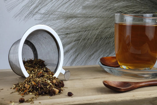 Rooibostee Sanddorn Sahne, loser Rooibostee im Teesieb, Teetasse mit bernsteinfarbenem Aufguss und Holzlöffel auf Holzuntergrund
