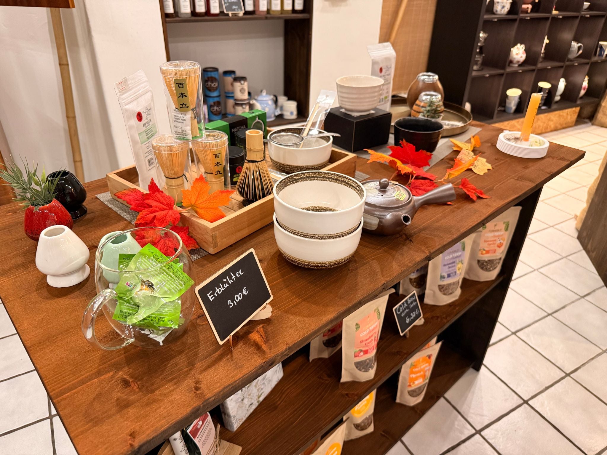 Tisch im Teegeschäft TeePunkt Erkelenz mit Matcha-Zubehör, Teeschalen, Teesieben und Teepackungen – herbstlich dekorierte Teepräsentation.