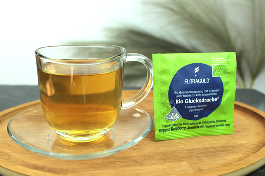 Produktfoto einer Tasse Tee und der Verpackung von Bio Glücksdrache, Teebeutel für sanften Genuss, Bio Grünteemischung mit Kräutern und Fruchtstücken, Himbeer-Jasmin-Geschmack.