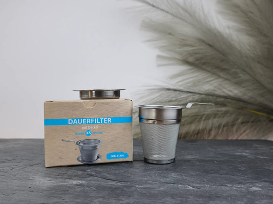 Produktfoto der Verpackung und des Dauerfilters mit Deckel in Größe XS (Ø 5 cm) aus Edelstahl, perfekt für die Teezubereitung im Becher oder Tea-for-One Set.