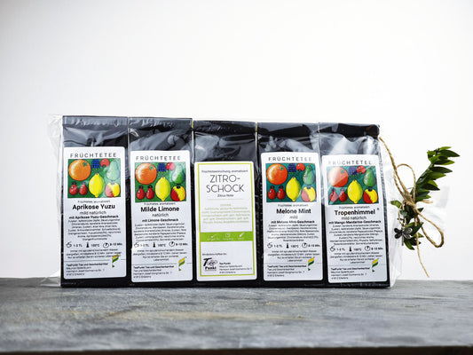 Fünf Packungen Früchtetee nebeneinander aufgereiht: Aprikose Yuzu, Milde Limone, Zitro-Schock, Melone Mint und Tropenhimmel, zusammengebunden mit einer Schleife
