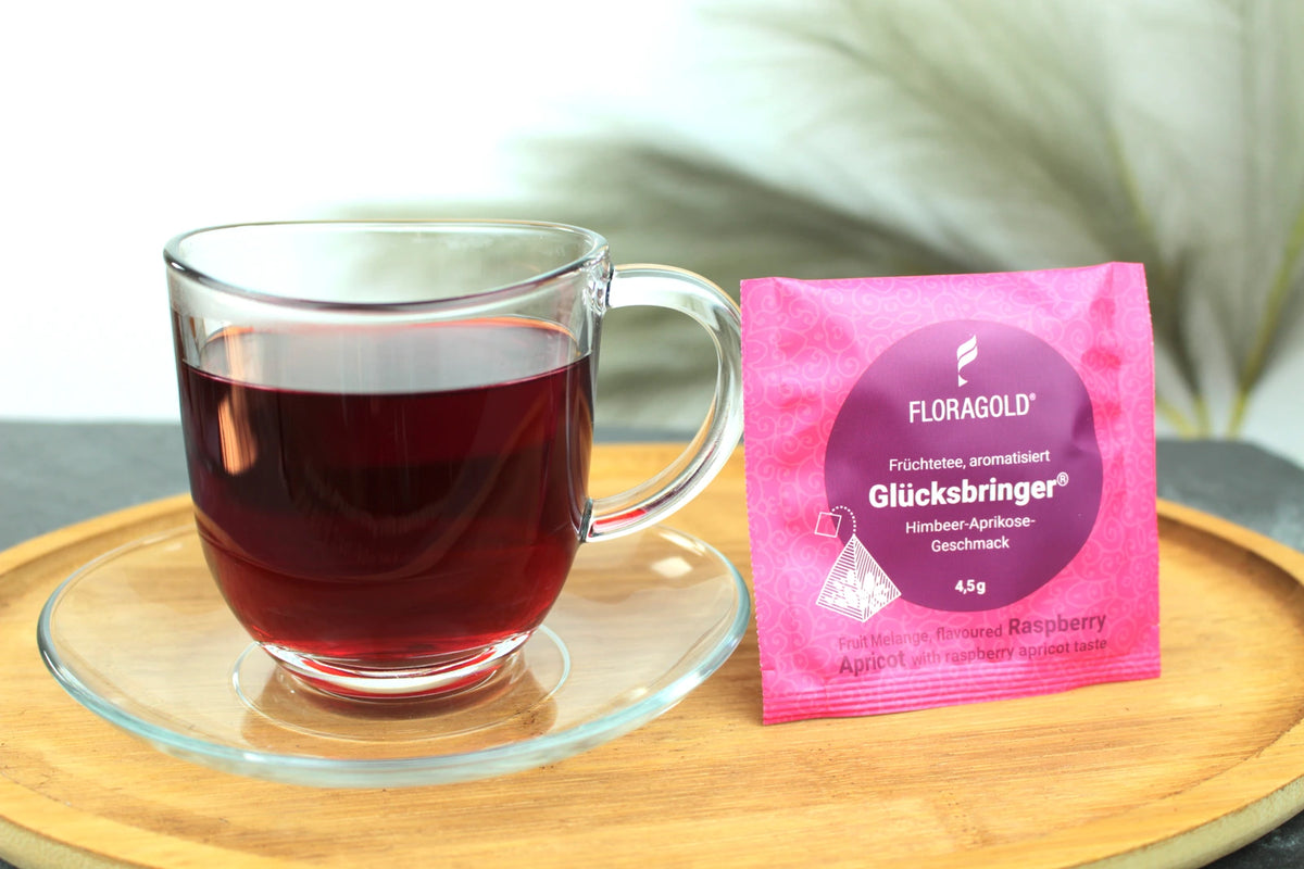 Produktfoto einer Tasse Tee und der Verpackung von Glücksbringer, fruchtiger Tee in Teebeuteln, Früchtetee mit Himbeer-Aprikose-Geschmack.