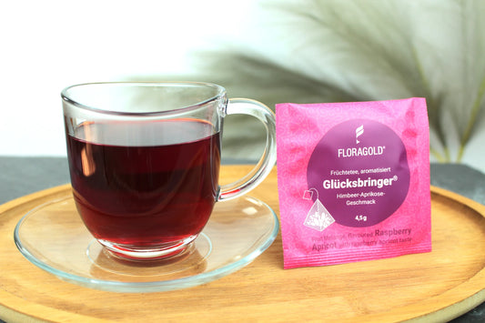 Produktfoto einer Tasse Tee und der Verpackung von Glücksbringer, fruchtiger Tee in Teebeuteln, Früchtetee mit Himbeer-Aprikose-Geschmack.