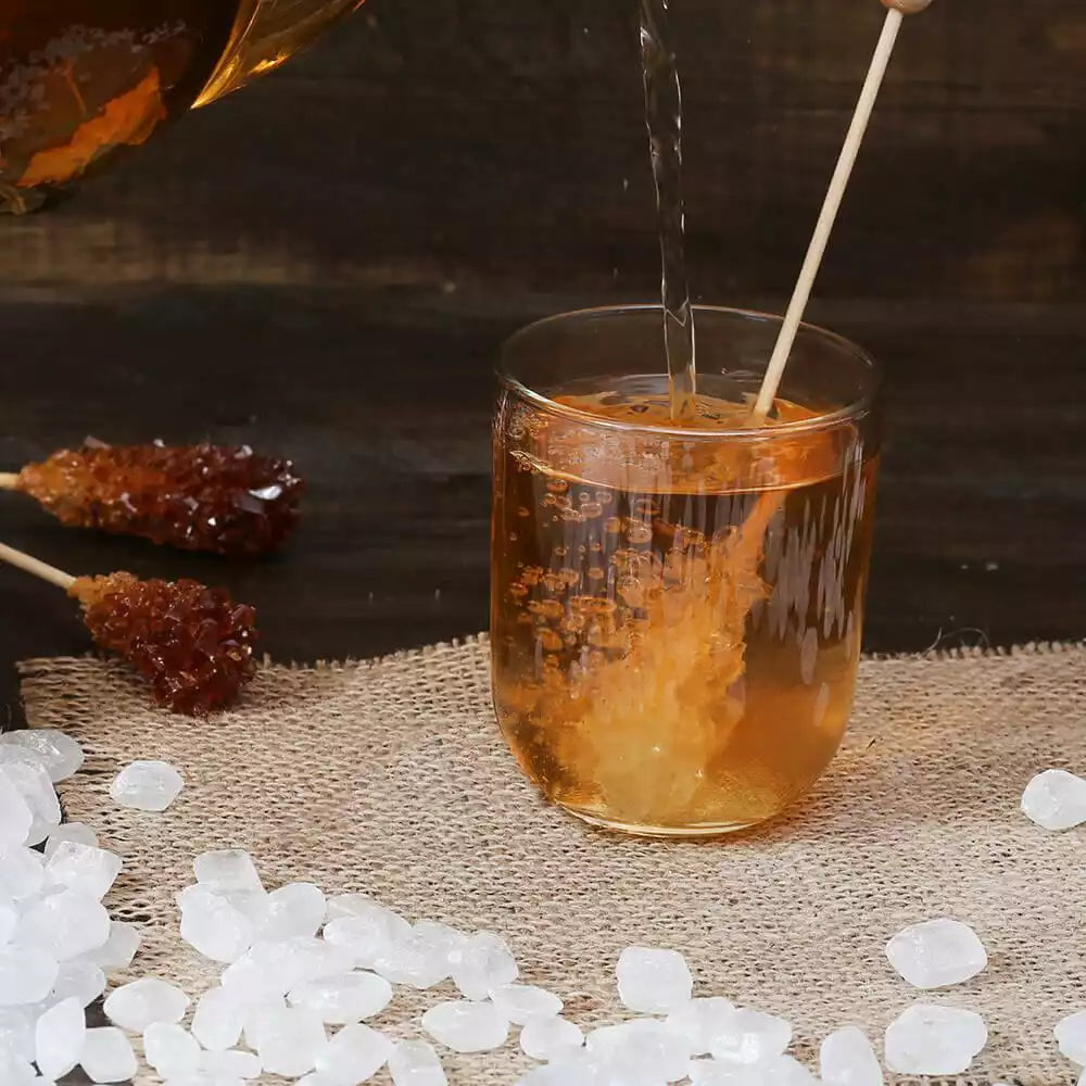Bernsteinfarbener Tee wird in ein Glas mit Kandiszucker-Stick gegossen, umgeben von losem Kandiszucker und Kandiszucker-Sticks auf einer Leinenmatte