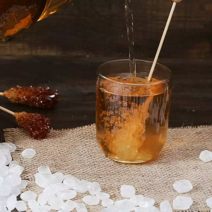 Bernsteinfarbener Tee wird in ein Glas mit Kandiszucker-Stick gegossen, umgeben von losem Kandiszucker und Kandiszucker-Sticks auf einer Leinenmatte