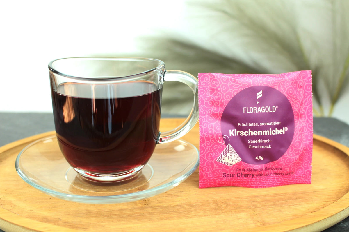 Produktfoto einer Tasse Tee und der Verpackung von Kirschenmichel, fruchtiger Teegenuss mit Sauerkirsch-Geschmack.