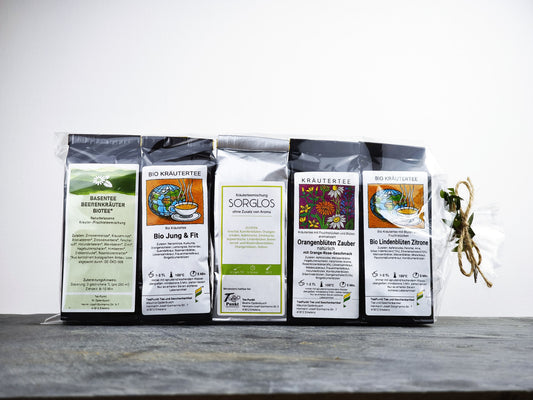 Fünf Packungen Kräutertee nebeneinander aufgereiht: Basentee Beerenkräuter, Bio Jung & Fit, Sorglos, Orangenblüten Zauber und Bio Lindenblüten Zitrone, zusammengebunden mit einer Schleife