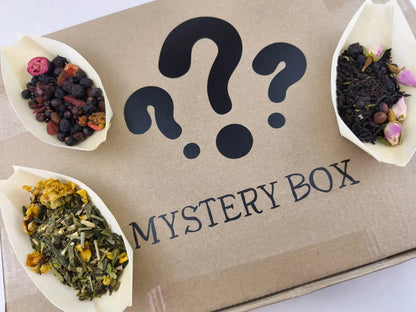 Karton mit der Aufschrift "Mystery Box" und Fragezeichen, umgeben von drei Papiertüten mit verschiedenen losen Teemischungen: Früchtetee mit dunklen Beeren, Schwarztee mit Rosenblüten und Kräutertee mit Ringelblumenblüten