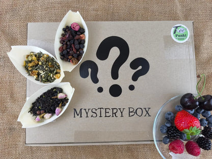 Karton mit der Aufschrift "Mystery Box" und Fragezeichen auf einer Leinenmatte, umgeben von drei losen Teemischungen (Früchtetee, Grüntee, Schwarztee mit Rosenblüten) und einer Schüssel frischer Beeren