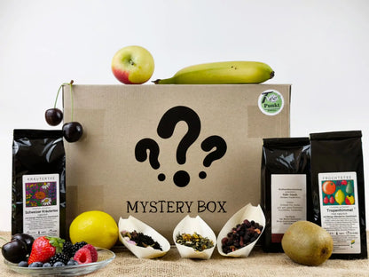 Karton mit der Aufschrift "Mystery Box" und Fragezeichen, umgeben von zwei Teepackungen (Kräutertee und Früchtetee "Tropenhimmel"), drei losen Teeproben und frischem Obst wie Apfel, Banane, Kirschen, Kiwi und Beeren
