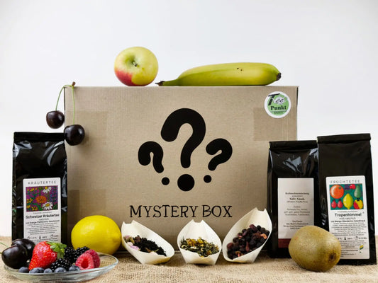 Karton mit der Aufschrift "Mystery Box" und Fragezeichen, umgeben von zwei Teepackungen (Kräutertee und Früchtetee "Tropenhimmel"), drei losen Teeproben und frischem Obst wie Apfel, Banane, Kirschen, Kiwi und Beeren
