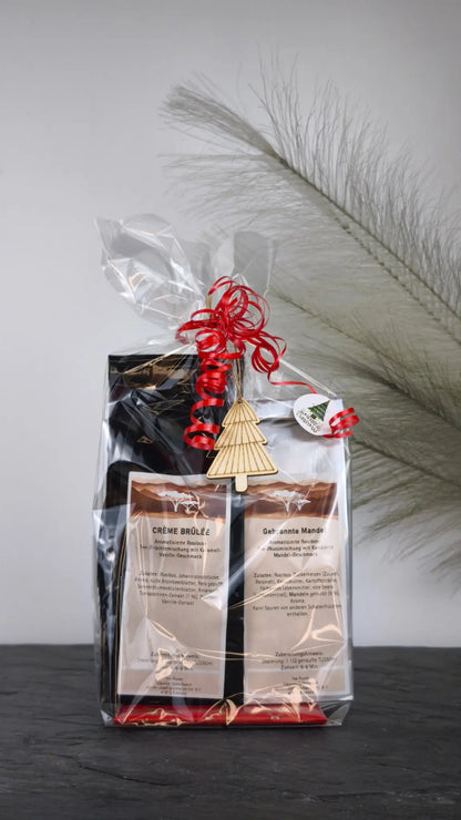 Geschenkset mit Crème Brûlée und Gebrannte Mandel Tee, festlich verpackt in Geschenkfolie mit Weihnachtsanhänger und roter Schleife.