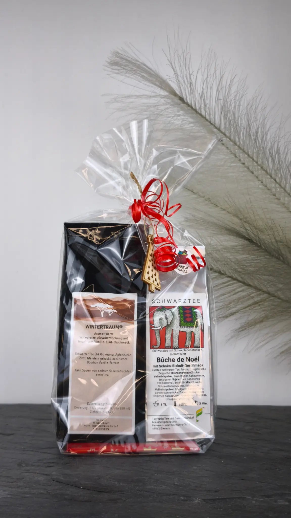Weihnachtliches Tee Geschenkset mit Wintertraum und Bûche de Noël, dekorativ verpackt in transparenter Folie mit rotem Band und Holzanhänger.