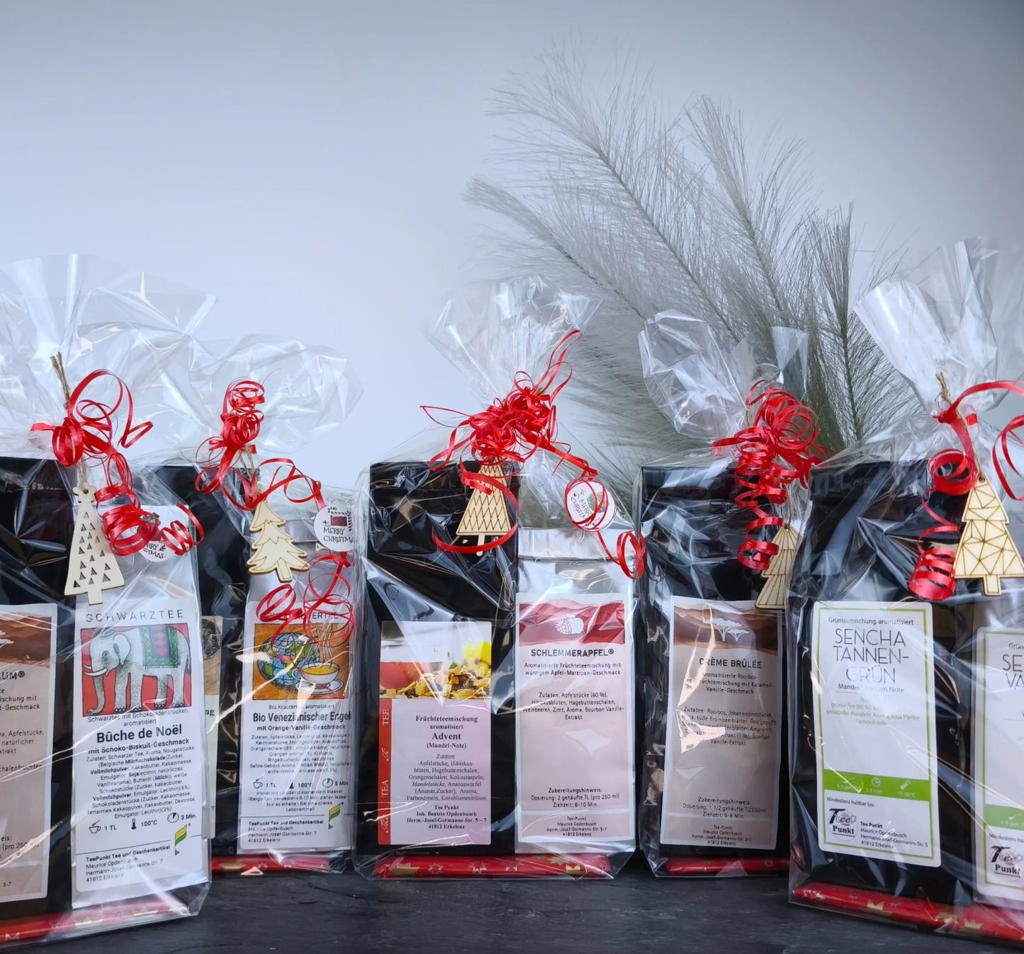 Weihnachtlich verpackte Tee-Geschenksets in transparenten Folientüten mit roten Schleifen und kleinen Holzanhängern, sortiert nebeneinander vor hellem Hintergrund.