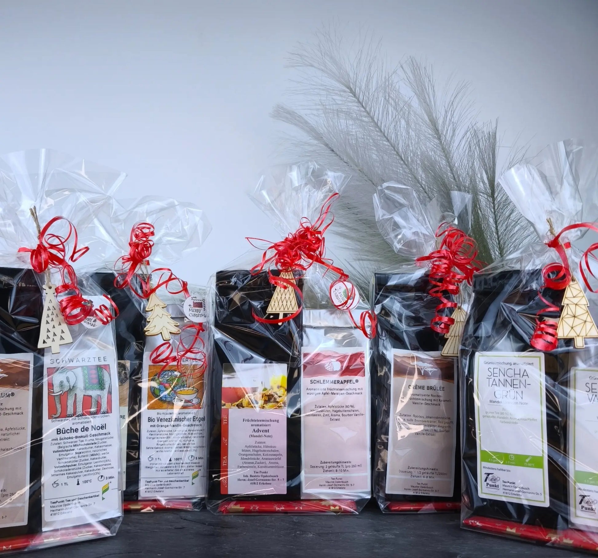 Weihnachtlich verpackte Tee-Geschenksets in transparenten Folientüten mit roten Schleifen und kleinen Holzanhängern, sortiert nebeneinander vor hellem Hintergrund.