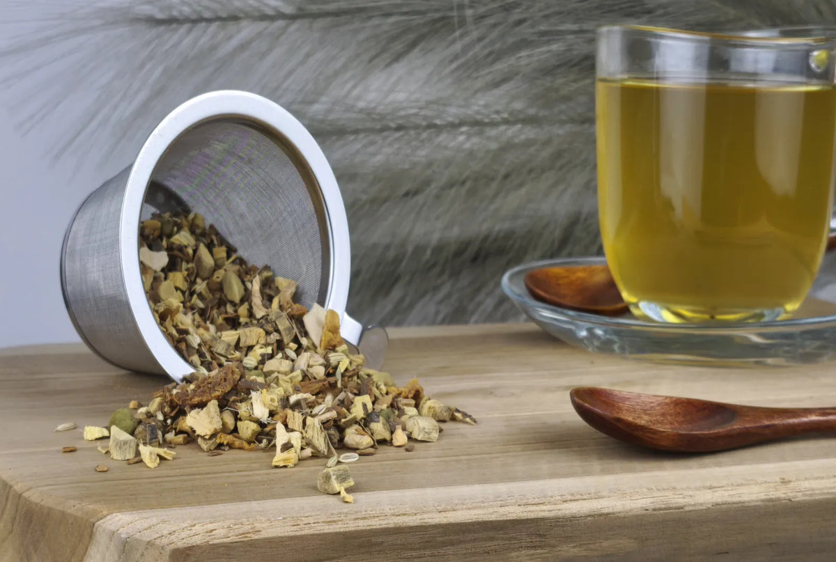 Loser Ayurveda-Tee "Vata" wird aus einem Sieb auf ein Holzbrett geschüttet, daneben eine Tasse goldgelber aufgebrühter Tee und ein Holzlöffel
