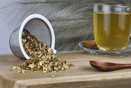 Loser Ayurveda-Tee "Vata" wird aus einem Sieb auf ein Holzbrett geschüttet, daneben eine Tasse goldgelber aufgebrühter Tee und ein Holzlöffel